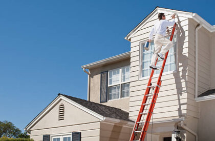 House painter with paint brush painting the trim of a 2nd story window of a Cape Cod Style house.